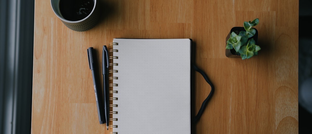 Warm wood desk flatlay with a notebook, pen, ceramic mug, and a sprig of leaves