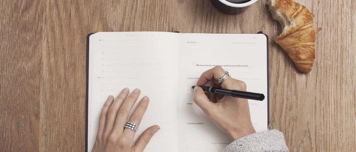 Hands writing in a notebook beside a mug of tea and a leafy plant on a warm wood table